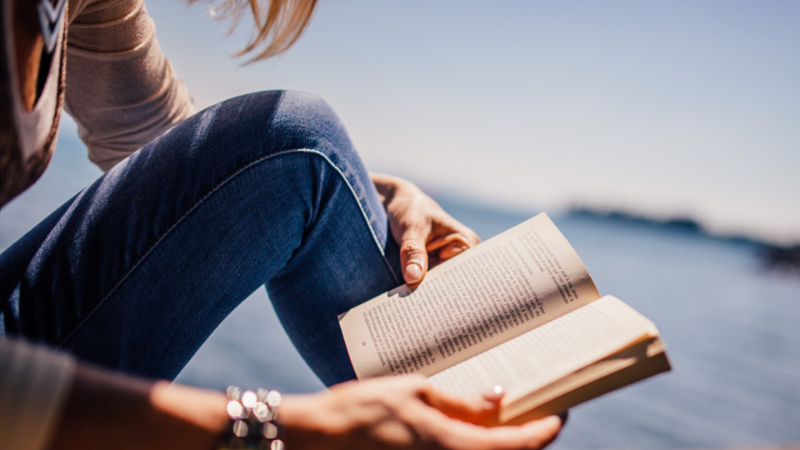 A woman reading a novel outside by a body of water on a sunny day.