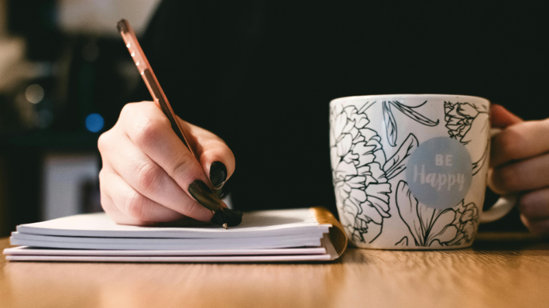 A closeup of a writer at work showing just her hand writing with a pen while she holds a cup of coffee in the other hand.